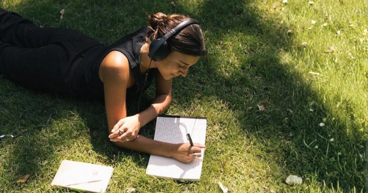 A woman laying on the grass in the park, looking thoughtful while holding a pen and writing for reflection.