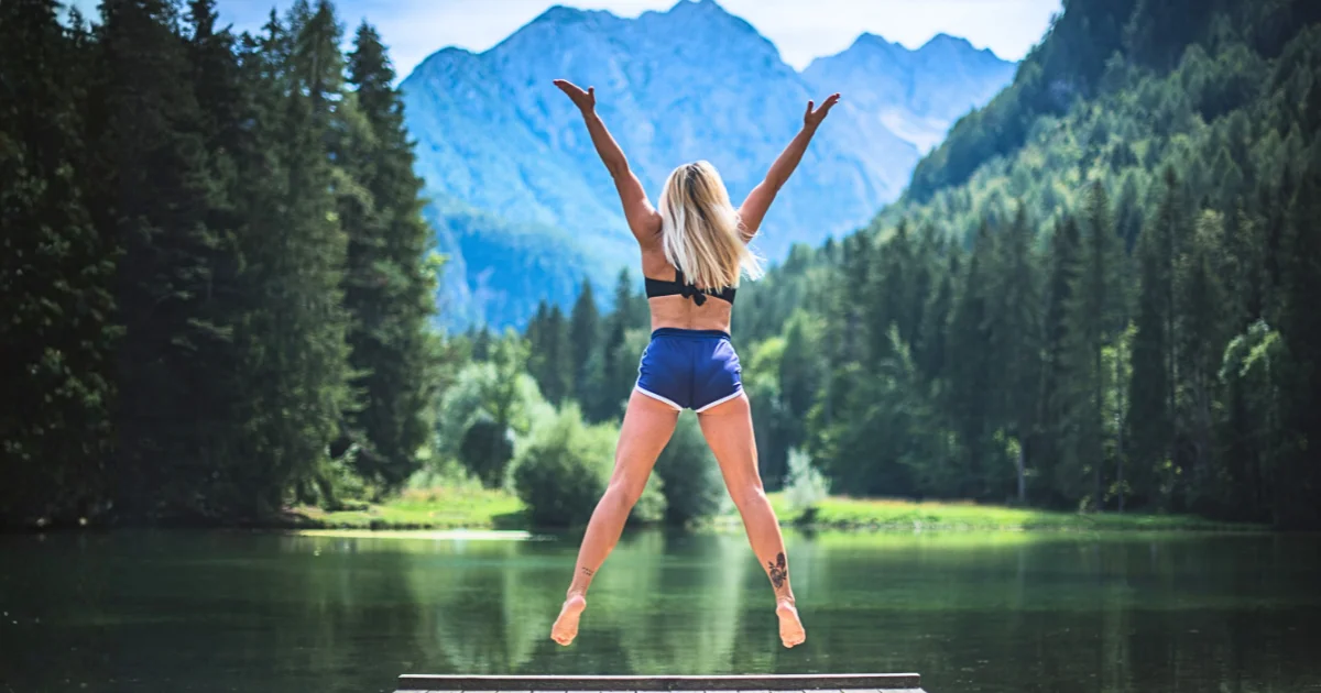 A person jumping with arms outstretched on a wooden dock overlooking a serene lake and mountains, illustrating natural ways to relieve stress through outdoor movement and exploration.